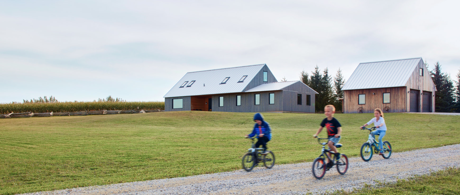 Three children riding bikes on a road by two wooden buildings and a field.